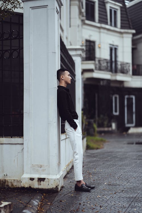 Side view full body of thoughtful young ethnic man in elegant attire with hands in pockets leaning on wall of aged building and looking away