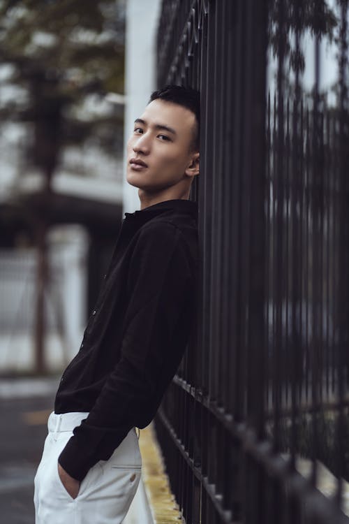 Side view of peaceful young Asian male in stylish clothes leaning on metal fence and looking at camera while standing in park with hands in pockets