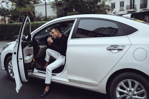 Side view of confident young Asian male millennial in smart casual outfit lighting cigarette while siting in modern car parked on asphalt road in city