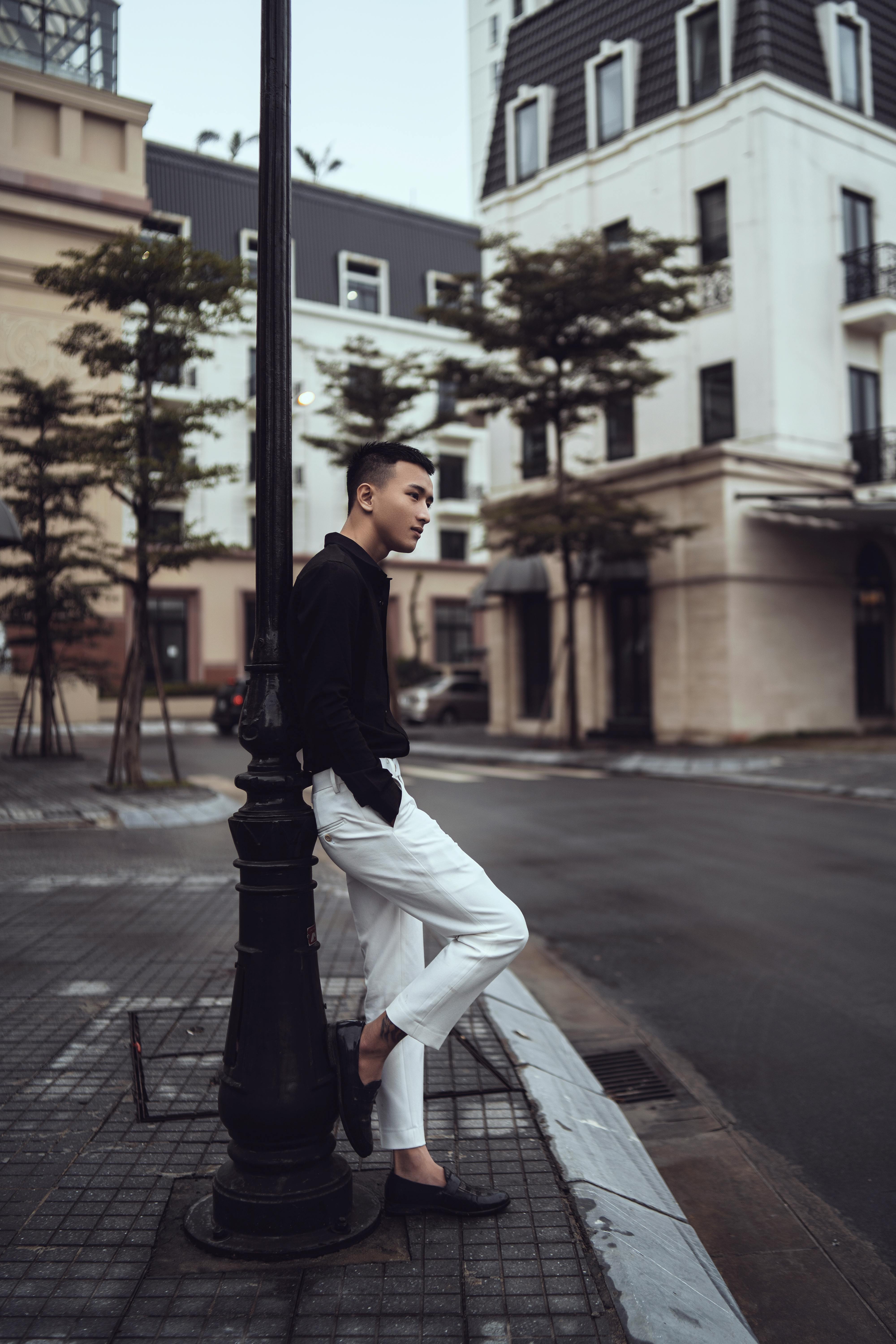 Side view of trendy young ethnic guy standing on paved sidewalk leaning on metal light pole and looking away near typical residential buildings on cloudy day