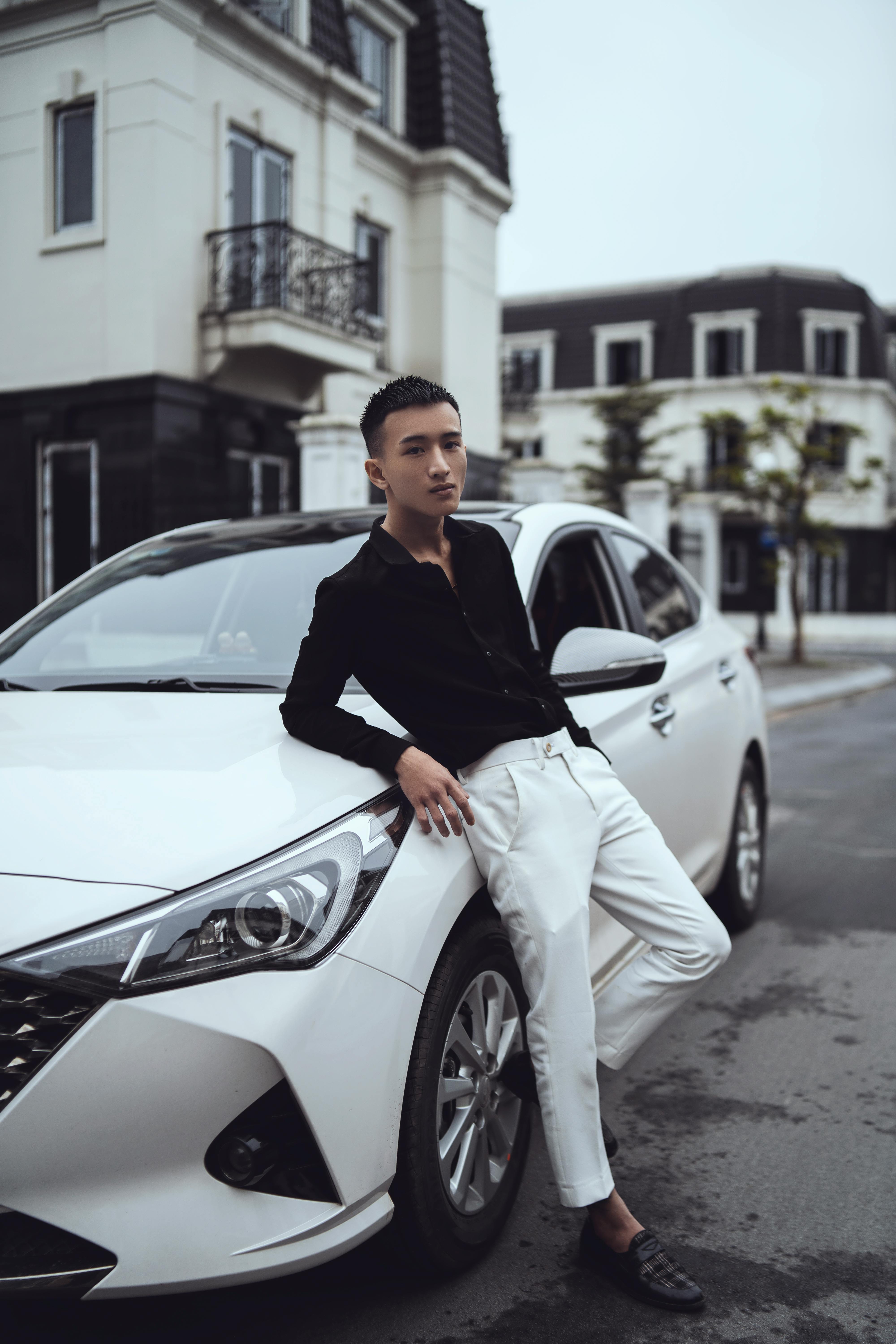 Full length of serious young ethnic man in fashionable outfit leaning on contemporary white car parked on phrasal road near residential house and looking at camera