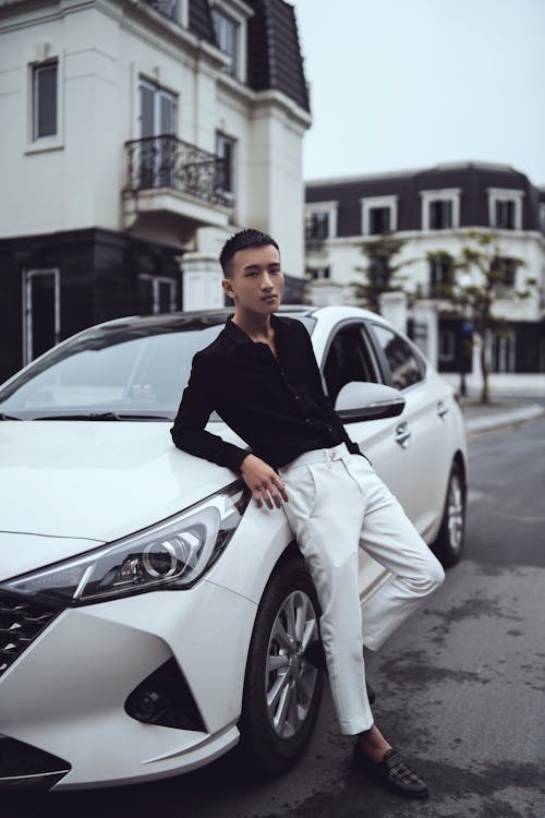 Self esteemed young Asian guy leaning on expensive car in city residential district