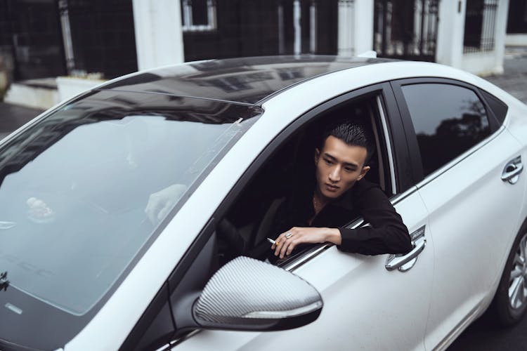 Stylish Young Ethnic Man Smoking Cigarette And Looking Out Window Of Modern Car