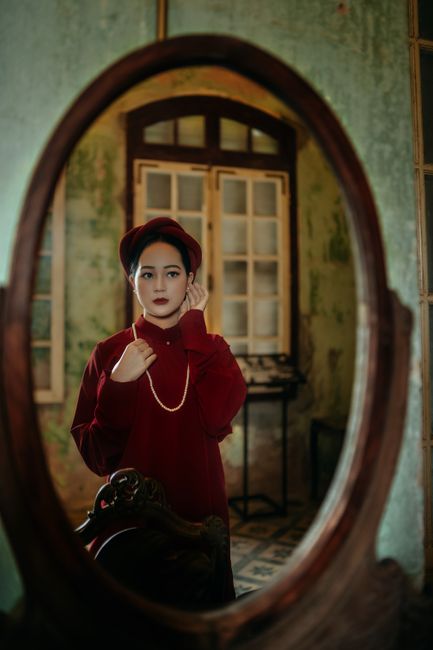 Contemplative young Asian female in elegant red dress and headgear looking in oval mirror in old cla