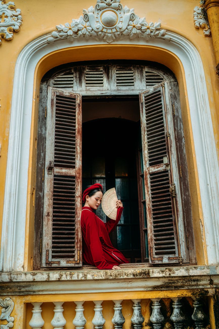 A woman in traditional red attire holding a fan sits in a vintage window frame.