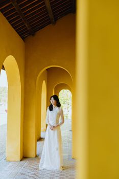 Full length young elegant Asian female in chic white dress standing in modern mansion archway with yellow walls and looking away in dreams