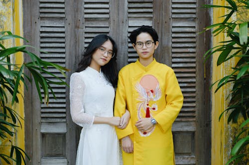Free Positive young Asian newlyweds wearing elegant authentic wedding clothes standing near mansion wooden doors and looking at camera Stock Photo