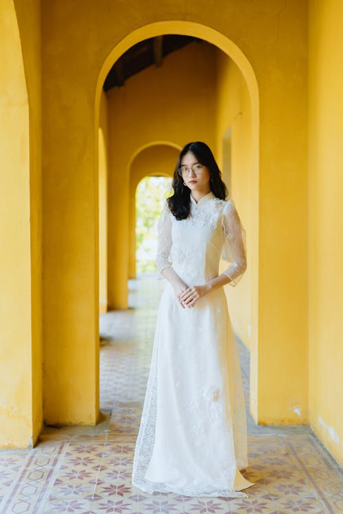 Full length calm young Asian female in classy maxi white dress standing in mansion archway and looking at camera