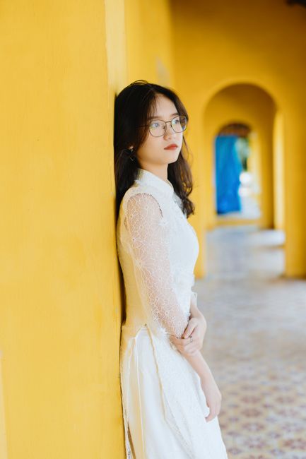 Side view elegant contemplative Asian female in white fancy dress standing in yellow building archway and looking away in dreams