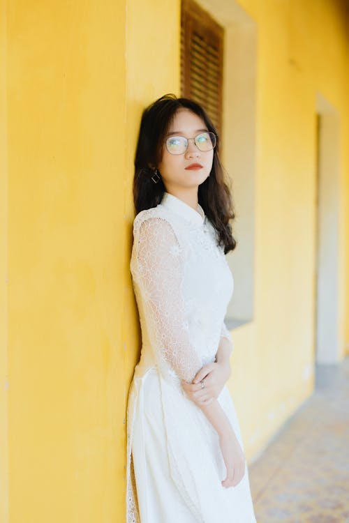 Side view unemotional young Asian bride wearing classy white dress leaning on yellow building wall and looking away in thoughts
