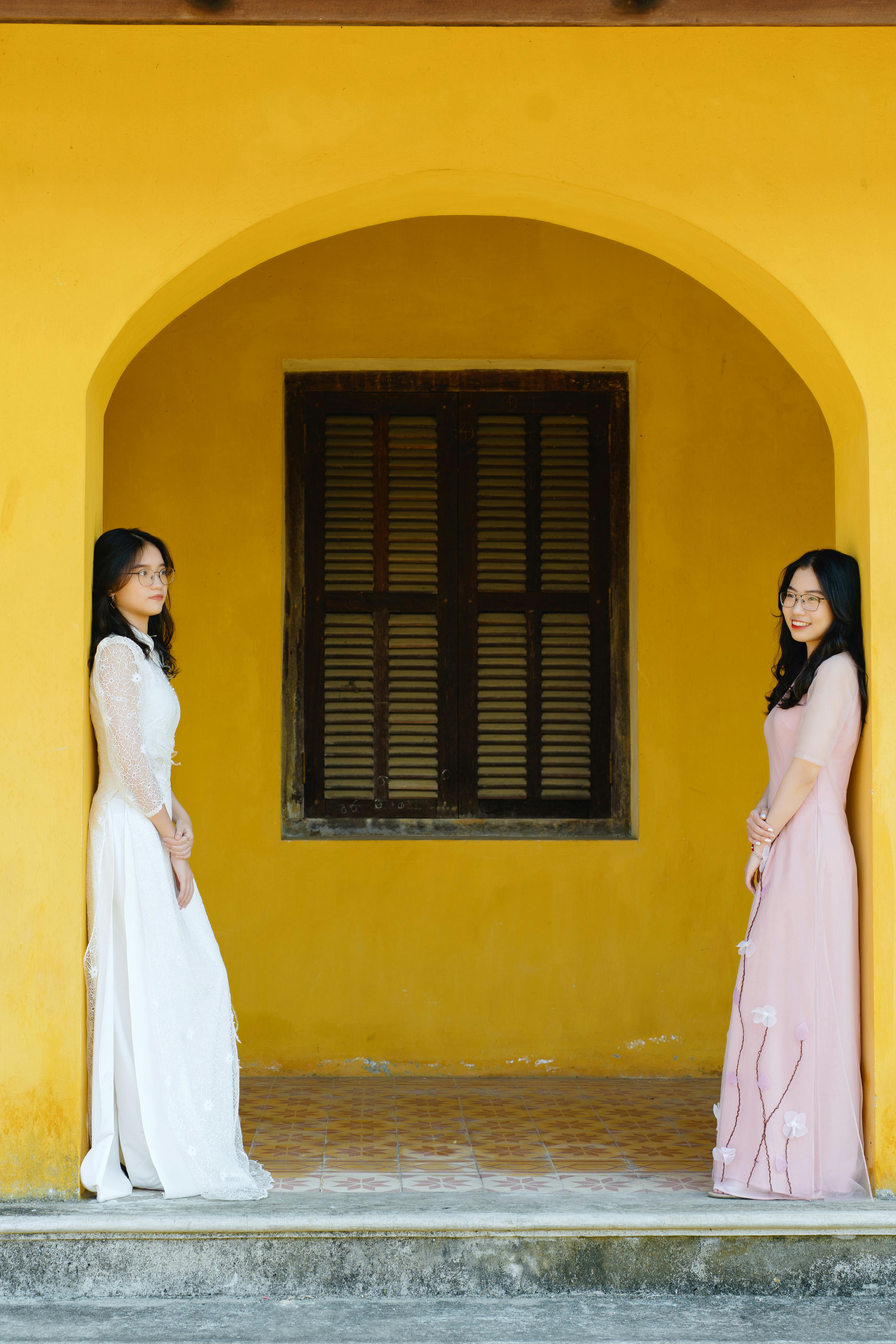 Two women in elegant dresses pose in a yellow archway, showcasing smiles and style.