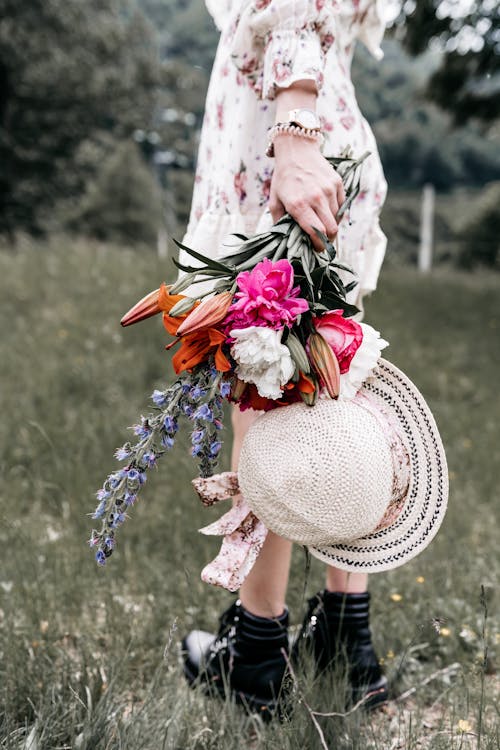 Back view of crop unrecognizable female in dress with hat and blossoming flowers standing on meadow