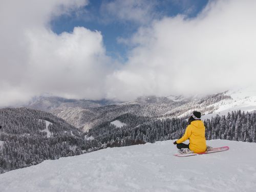 Free Snowboarder in yellow jacket sitting on snow, admiring a snowy mountain landscape under a cloudy sky. Stock Photo