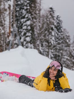 Smiling woman in a yellow jacket lying on snow with snowboard, enjoying winter outdoors.
