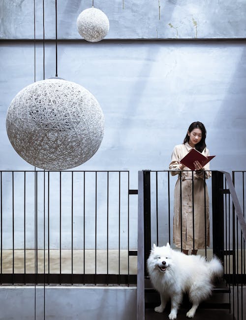 A Woman in a Beige Coat Reading a Book While Standing In Front of a White Dog