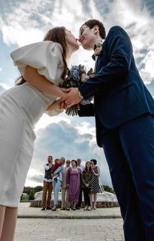 Bride and groom share a kiss with family witnessing. Captures love and togetherness outdoors.
