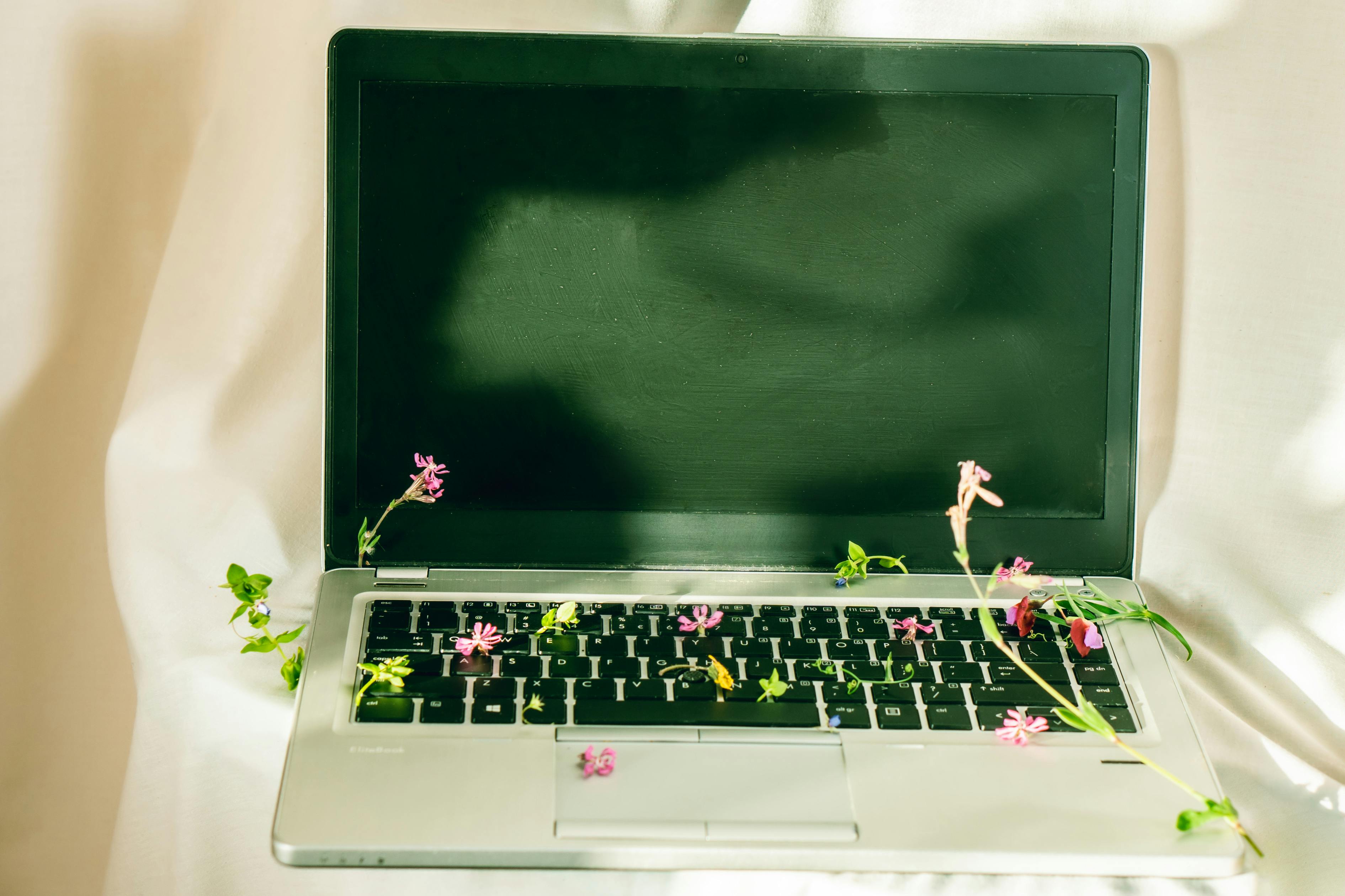 A silver laptop adorned with green vines and pink flowers on a beige studio background, representing nature and technology.