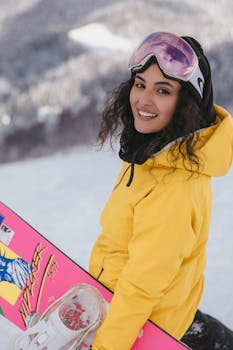 Smiling woman in yellow jacket with pink snowboard on a snowy mountain.
