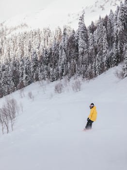 A snowboarder in a yellow jacket glides down a snow-covered mountain with a forest backdrop.