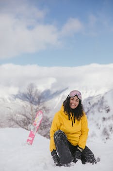 Woman in yellow jacket smiling in the snow with a snowboard and mountain view.