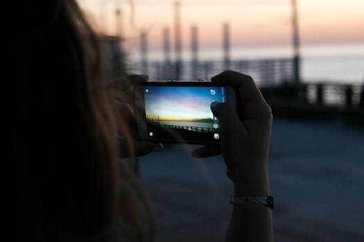 A person photographs a vibrant sunset with a smartphone by the waterfront. Outdoor scene.