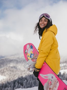 Smiling woman in yellow jacket holding a pink snowboard on a snowy mountain.