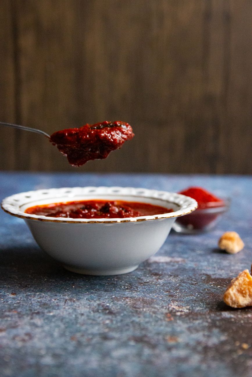 A small bowl filled with thick, glossy dark red-orange Ghanaian shito pepper sauce, with a wooden spoon resting alongside
