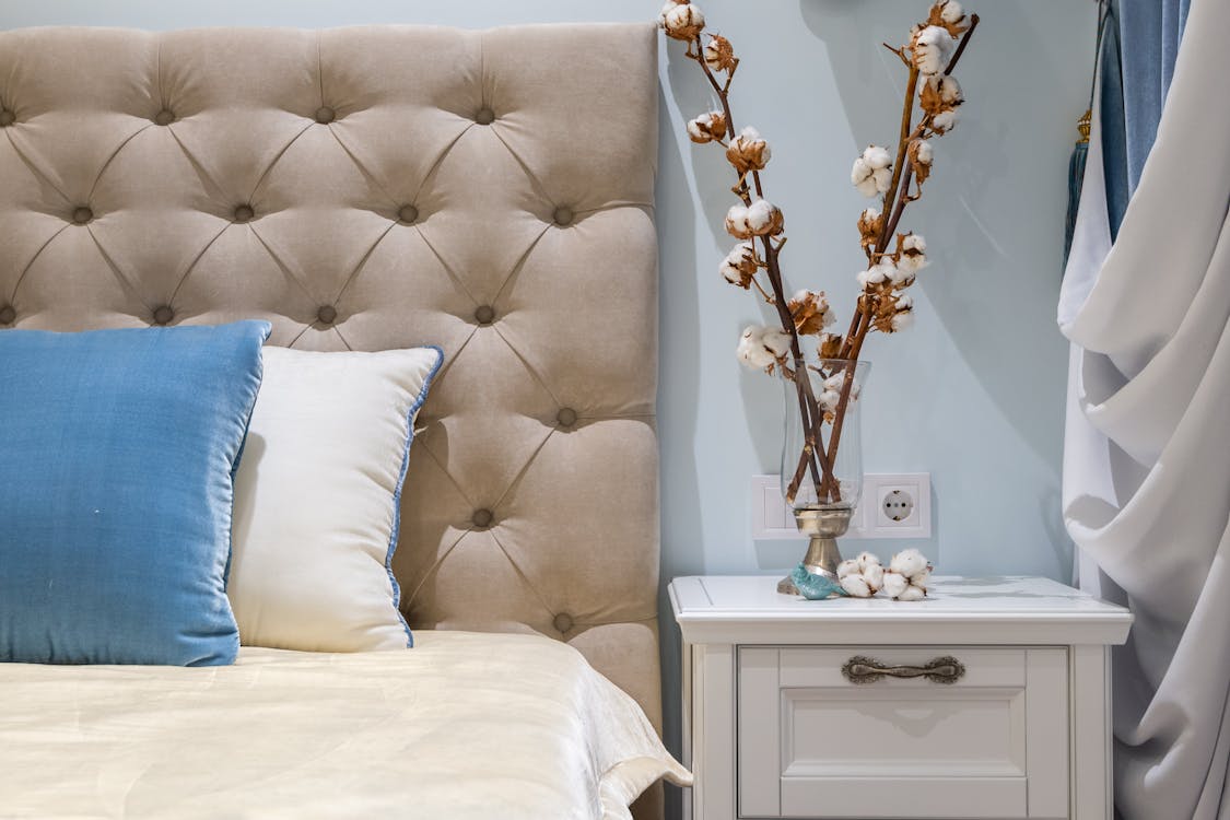 Cotton plants in vase placed on white nightstand near comfortable bed with soft cushions and headboard in classic styled bedroom