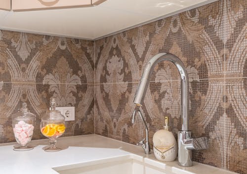 Contemporary kitchen stone counter with undercounter sink and shiny metal faucets at beige and brown tiled backsplash with ornament