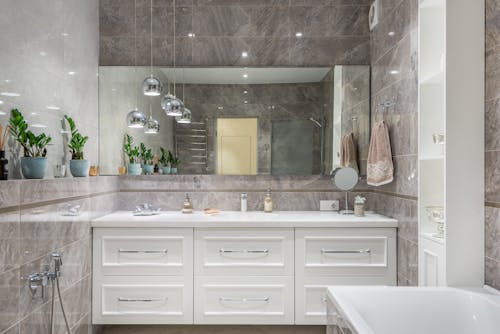 Interior of contemporary bathroom with gray marble tile and big mirror above white counter with washbasin in luminous lights
