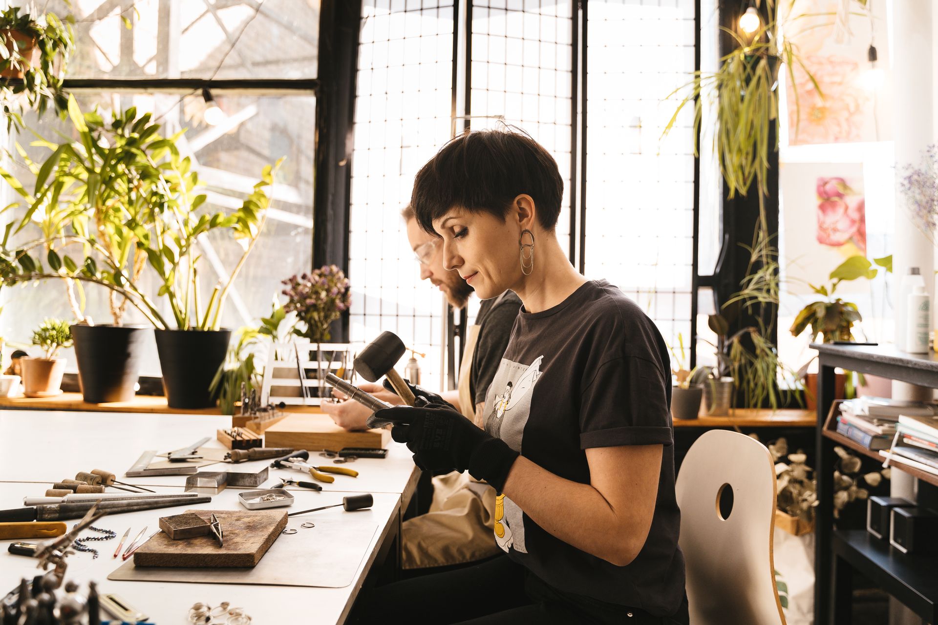 Two artisans working diligently at a crafts table in a sunlit, plant-filled workshop.