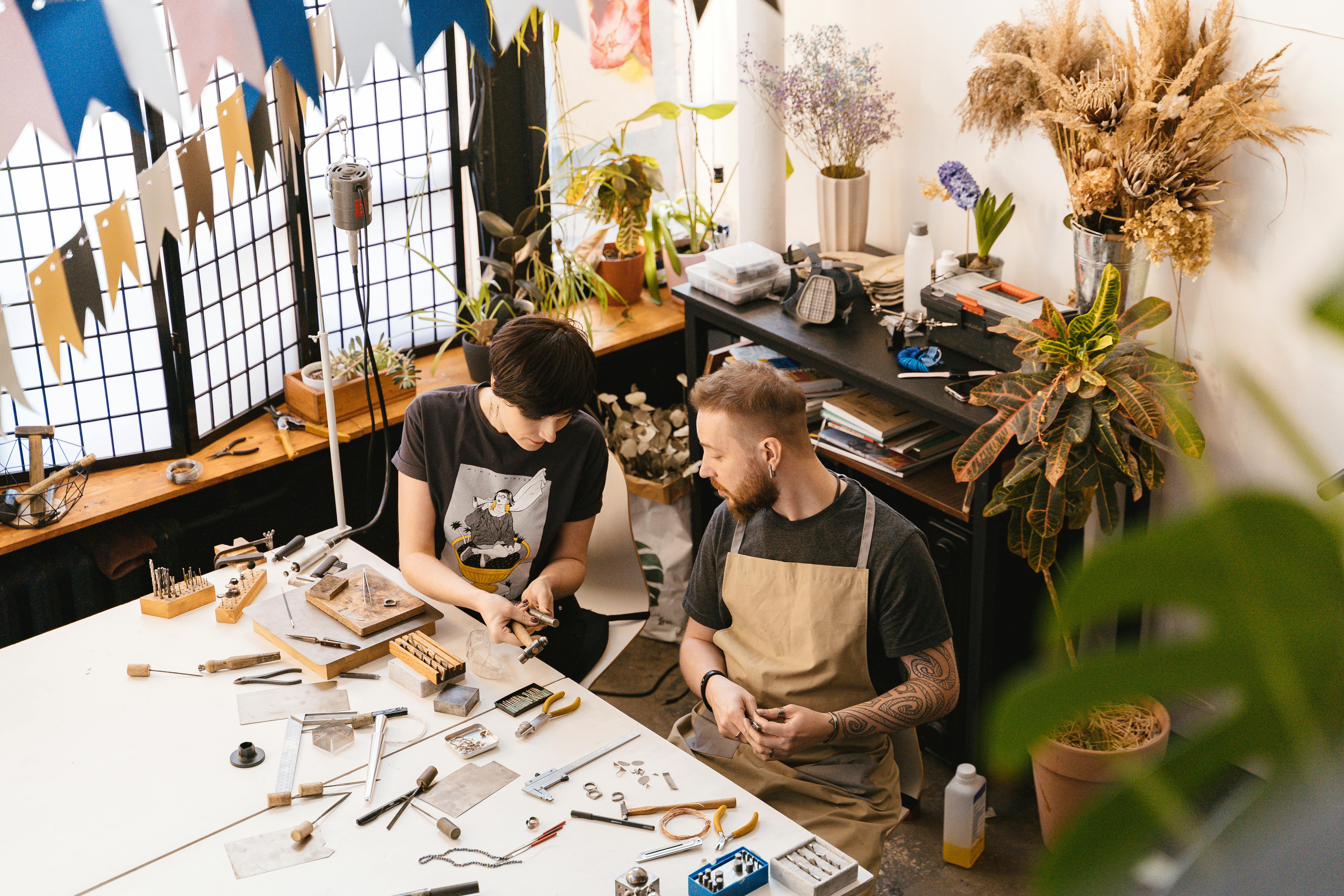 Two artisans creating jewelry pieces in a sunlit studio filled with plants and tools.