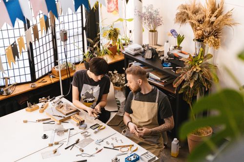 Free Two artisans creating jewelry pieces in a sunlit studio filled with plants and tools. Stock Photo