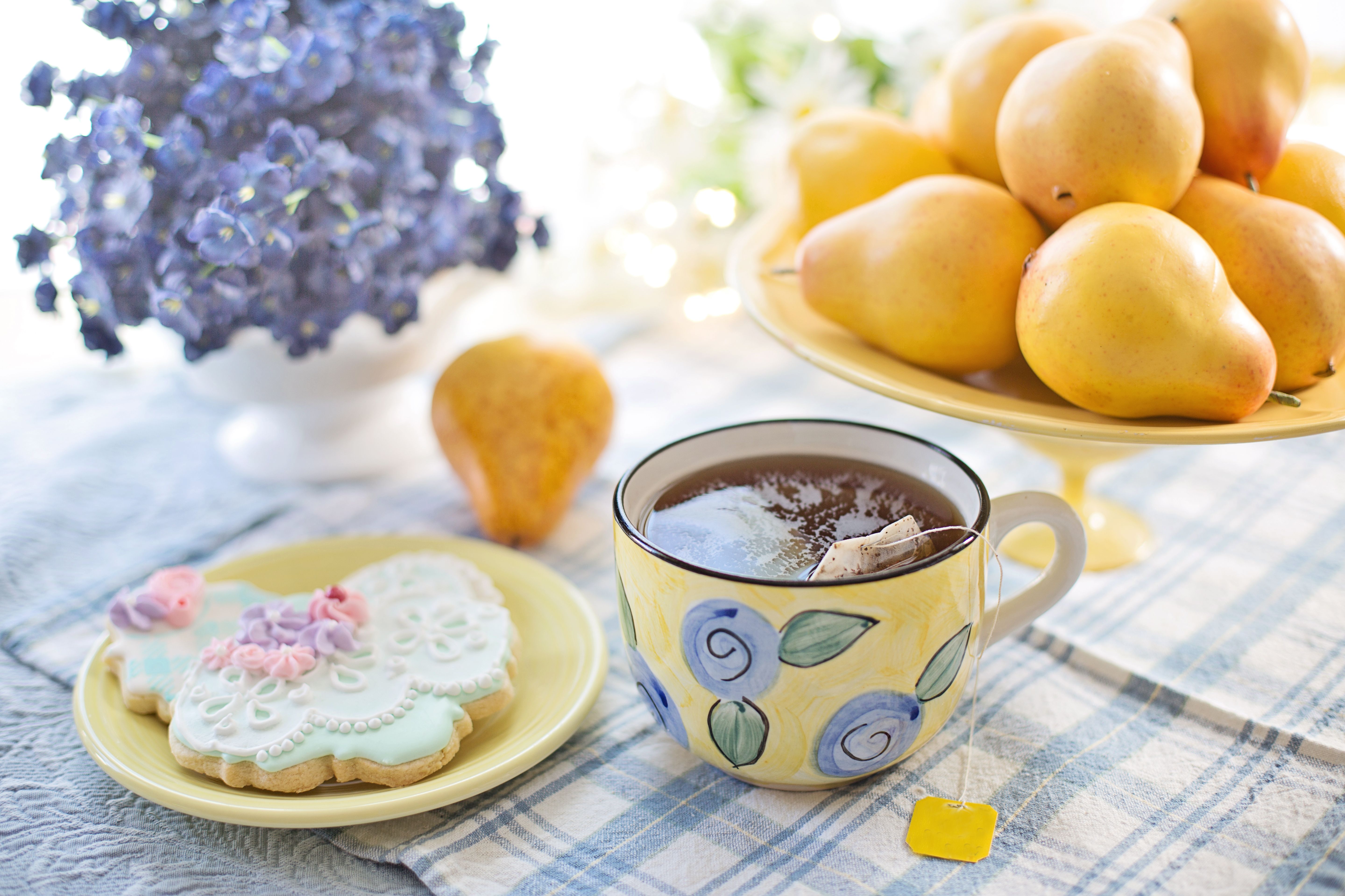 A charming tea setup with a floral cup, cookies, pears, and flowers on a checkered tablecloth.