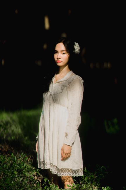 Elegant woman in a white dress stands amidst lush greenery, illuminated by soft sunlight.