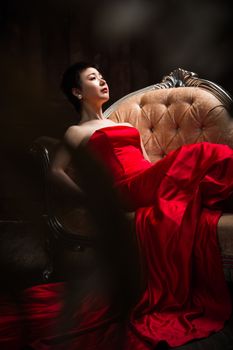 Sophisticated woman in a vibrant red gown reclining on an elegant antique chair indoors.