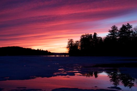 A vibrant sunset with colorful skies reflected over a frozen lake, framed by silhouette trees.
