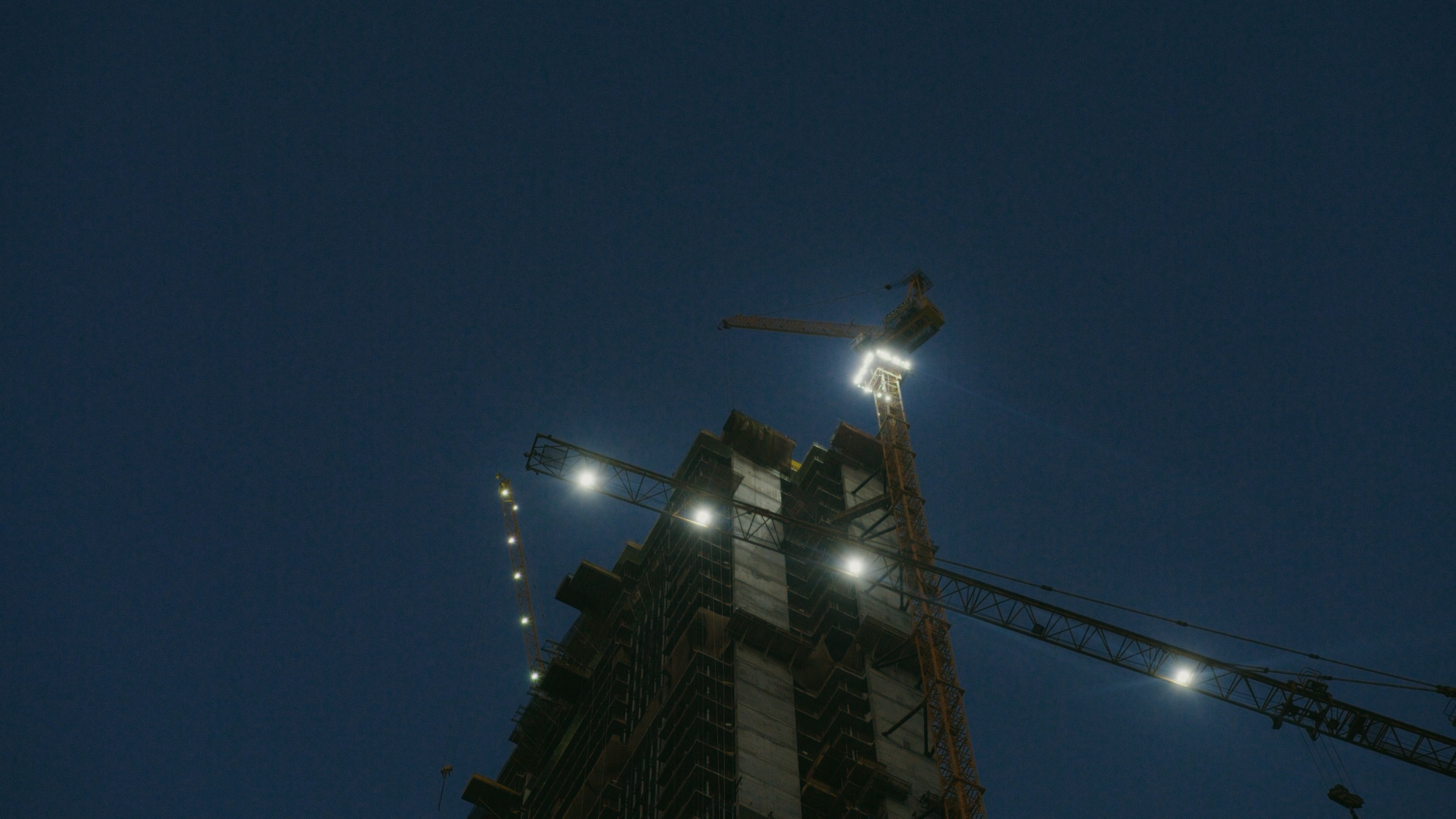 Illuminated tower cranes at a construction site captured during nighttime.