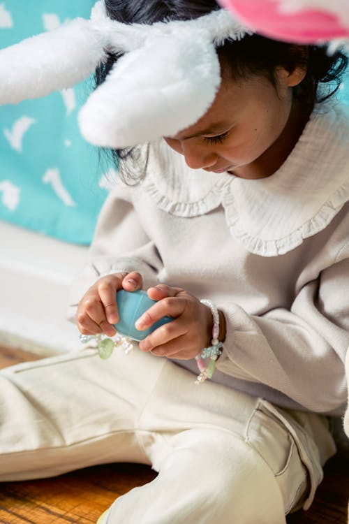 Free Crop ethnic child in hairband with decorative rabbit ears and painted egg sitting on floor on Easter Day Stock Photo