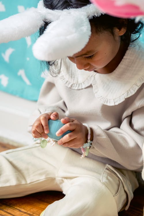 Crop ethnic child in hairband with decorative rabbit ears and painted egg sitting on floor on Easter Day