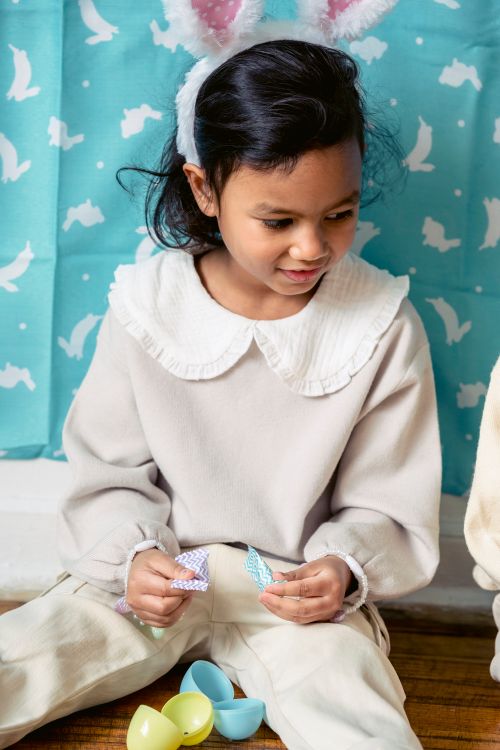 Free Ethnic child in hairband with decorative bunny ears sitting on parquet with ornamental paper pieces while looking down during festive event Stock Photo