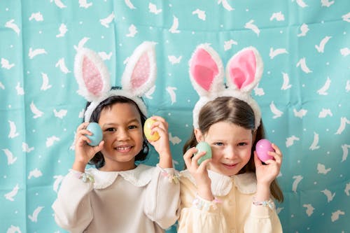 Free Content diverse children in hairbands with bunny ears showing decorative eggs while looking at camera against fabric with rabbit ornament Stock Photo