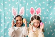 Content diverse children in hairbands with bunny ears showing decorative eggs while looking at camera against fabric with rabbit ornament