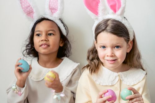Free Cute content multiracial girls in light wear and funny bunny ears playing with multicolored toy Easter eggs against white wall Stock Photo
