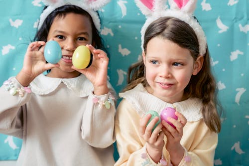 Happy multiethnic girls in light clothes and bunny ears playing with multicolored toy Easter eggs against blue background in playroom