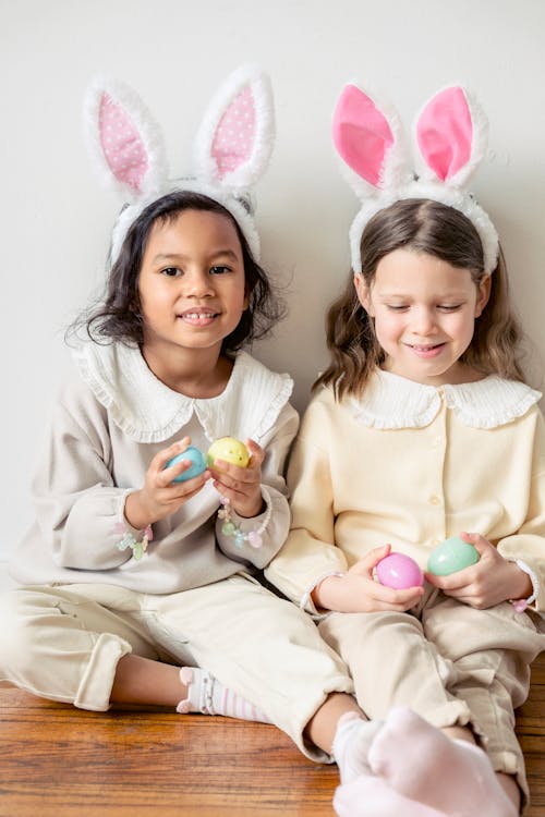 Adorable diverse girls wearing soft light clothes and funny bunny ears sitting on floor together with toy eggs