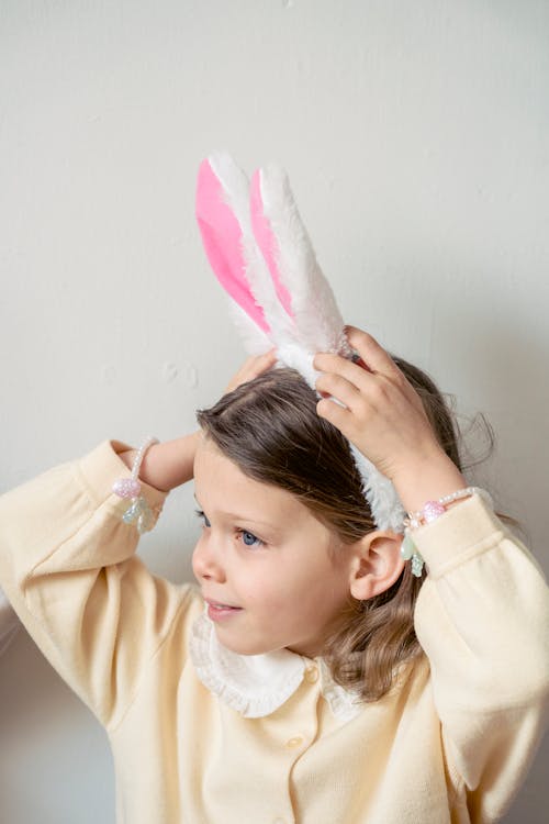 Cute little girl in light dress putting on funny bunny ears against white wall and looking away