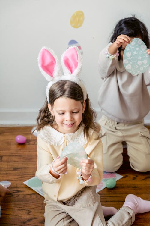 Free Adorable little girls wearing bunny ears playing with blue decorative Easter stickers while sitting on floor Stock Photo