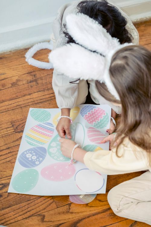 Free From above of unrecognizable little girls with fluffy bunny ears headband witting on wooden floor and playing with colorful egg shaped stickers during Easter celebration Stock Photo