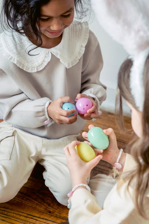 Free From above of crop cute happy diverse little girls in stylish clothes playing with colorful Easter egg toys while sitting on wooden floor at home Stock Photo